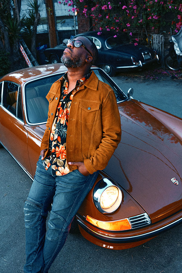 A man in a brown jacket, patterned shirt and jeans leans against a luxury sportscar