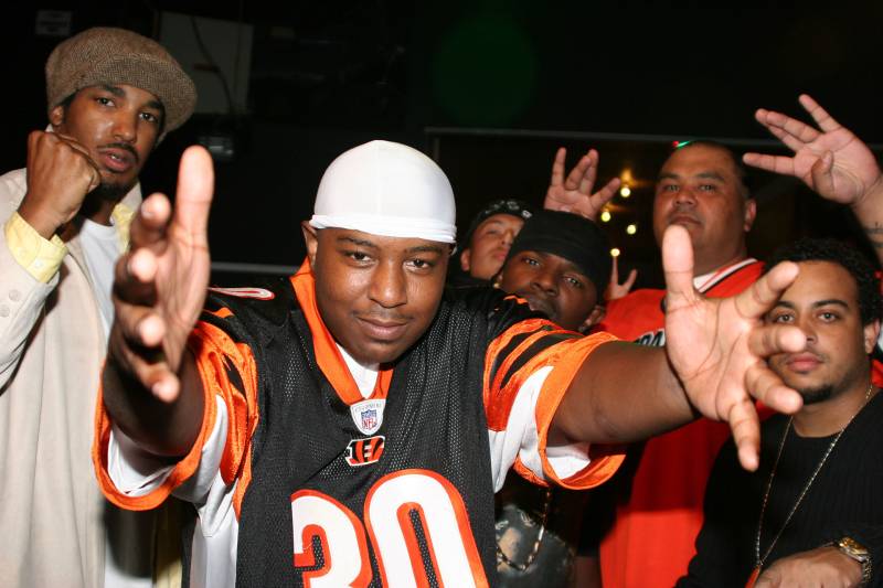 A man in a durag and football jersey holds his arms spread, with friends in the background