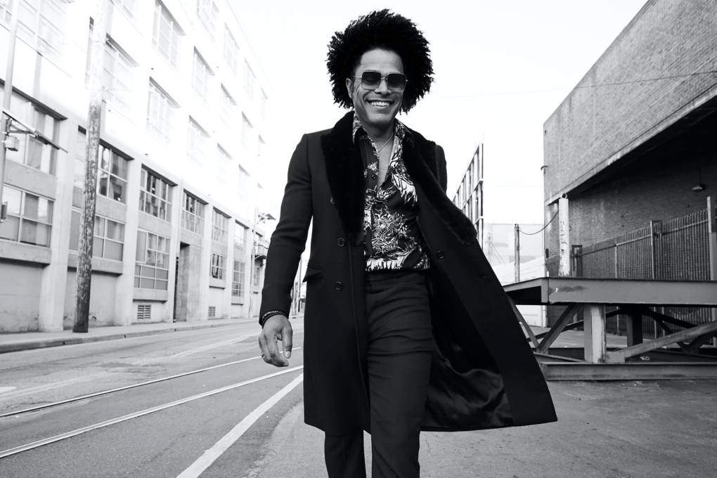 a tall Black man wearing sunglasses and a long coat smiles while walking down a city street in a black and white photograph
