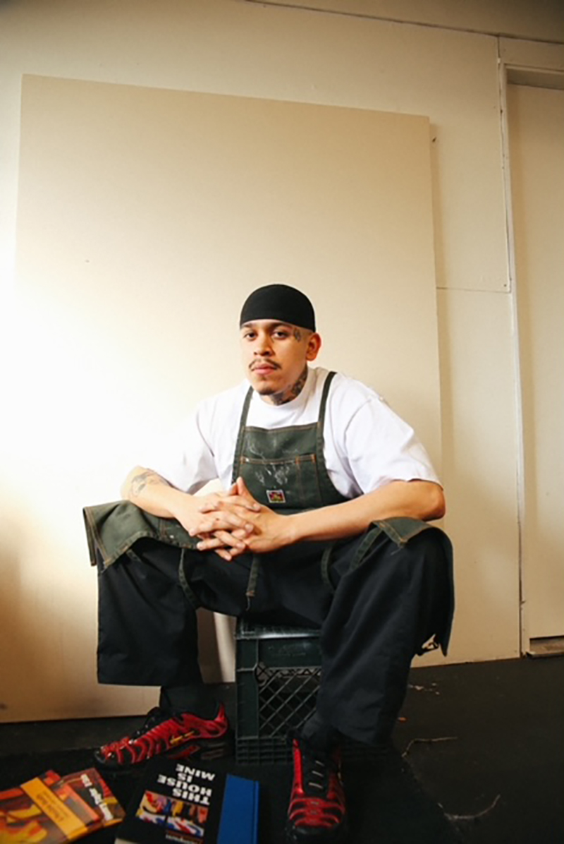 Young man in work apron and black skullcap sits with hands clasped in front of a blank canvas