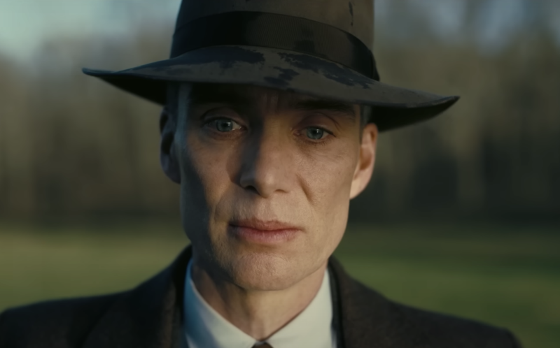 A thin white man with sharp cheekbones stands alone outside, concern etched on his face. He is wearing a brown suit and hat.