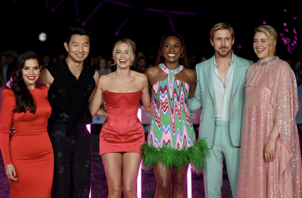 A Latina in a striking red dress, an attractive Asian man, a blond white woman, a smiling Black woman, a white man in a mint green suit and a white woman in a pink sequined cape and dress stand together in a line on a red carpet.