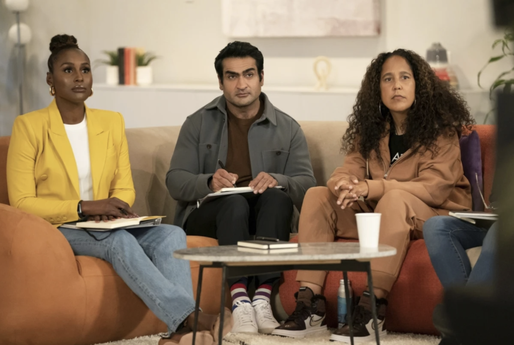 A man of South Asian descent sits on a couch between two Black women. All three are listening intently to someone off camera. The room has the look of a minimalist white living room.