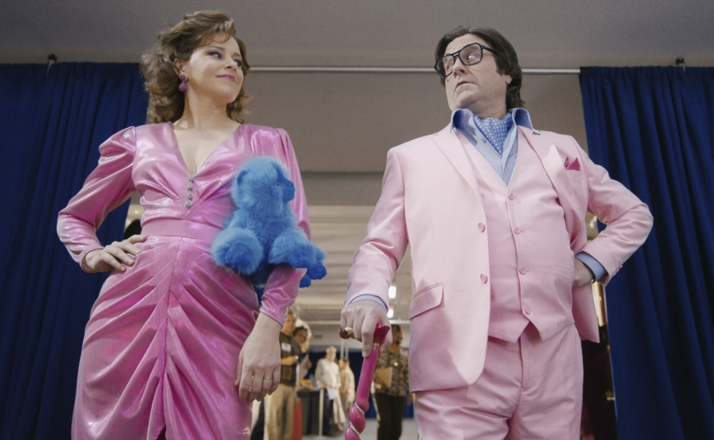 A woman in a tight fitting pink, pleated velvet dress clutches a soft blue toy and looks to her left at an older man wearing a pink three-piece suit and large glasses.