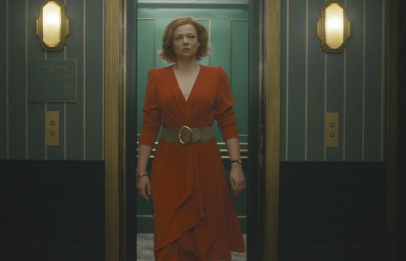An attractive woman with chin-length red hair walks through a doorway in a dark hall. She looks concerned.