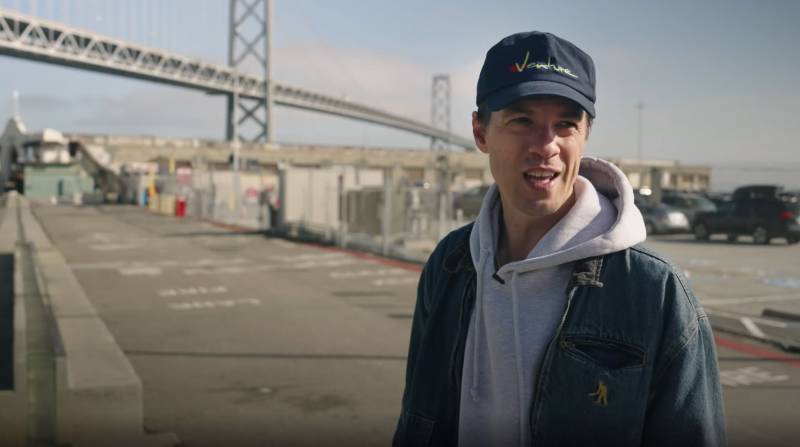 A man in a denim jacket and cap talks to the camera in front of the Bay Bridge