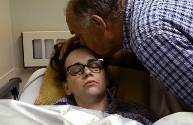 A young woman wearing glasses lies in a hospital bed, visibly in pain, while an older man strokes her hair and kisses her forehead.