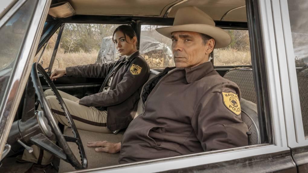 Two Indigenous police officers - one male, one female - sit in a car, wearing brown and tan uniforms. The environment outside the car's windows is desert.