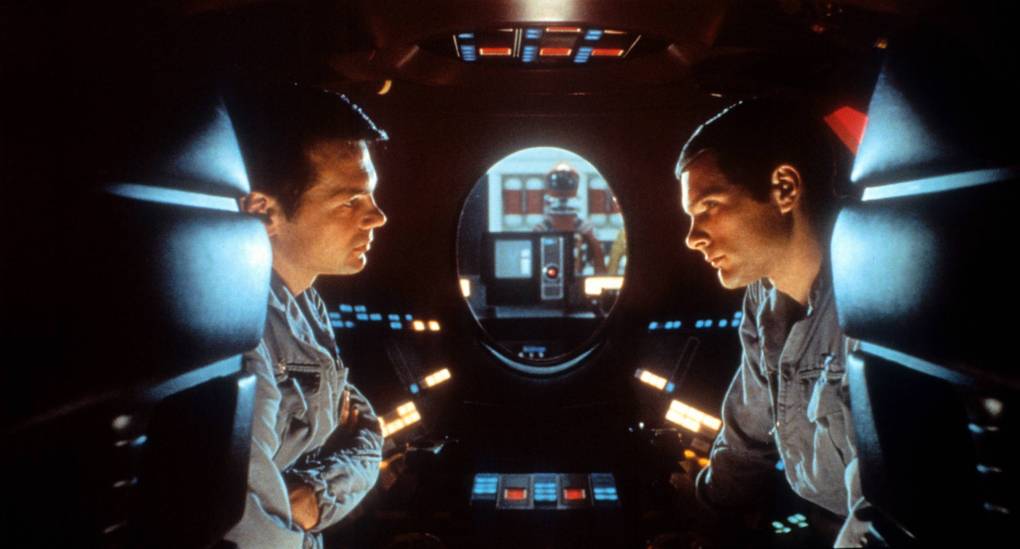 Two astronauts sit face to face in a small craft. Through an oval window between them, a robot is visible.