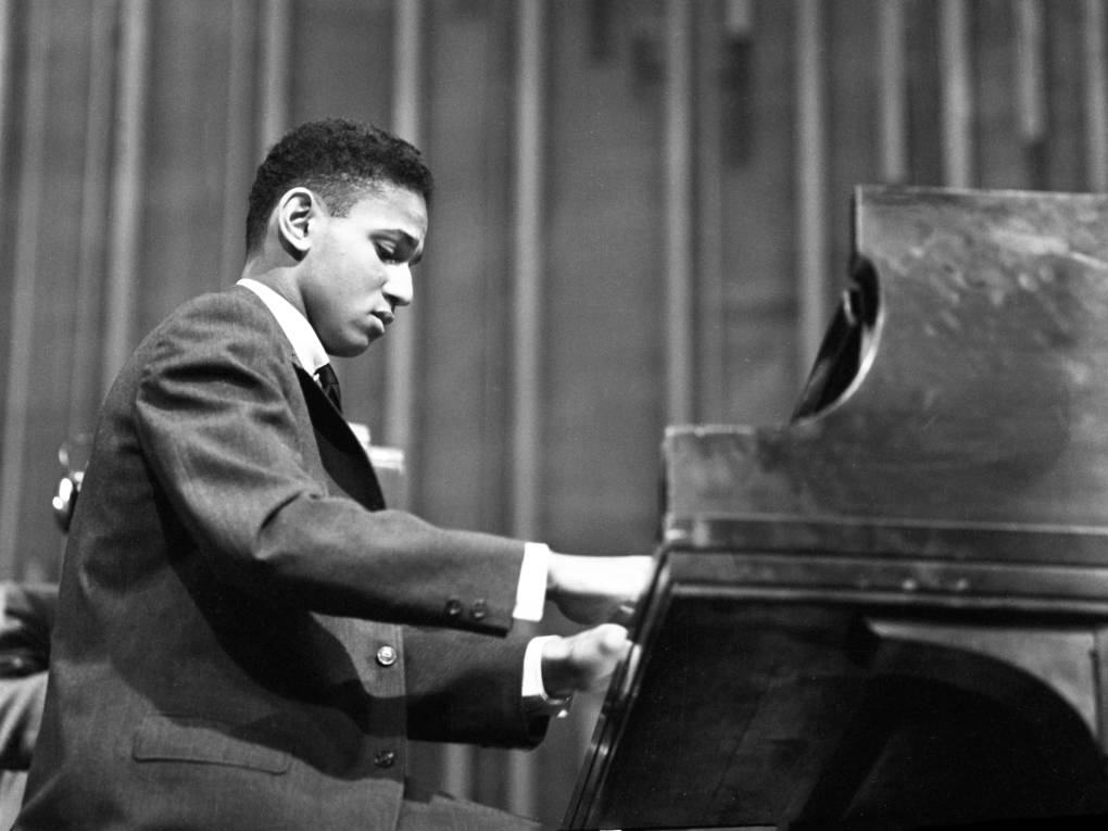 A young Black man with short hair in a suit plays the piano