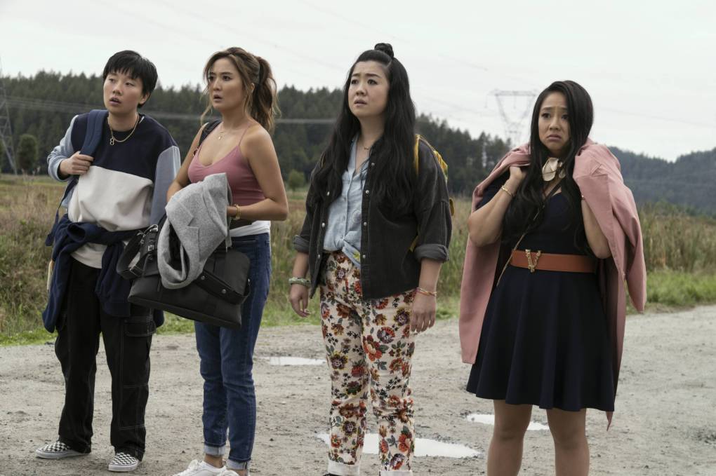 A nonbinary Asian person stands in the middle of a battered road alongside three Asian women. All four of them look very worried about what they see ahead of them.