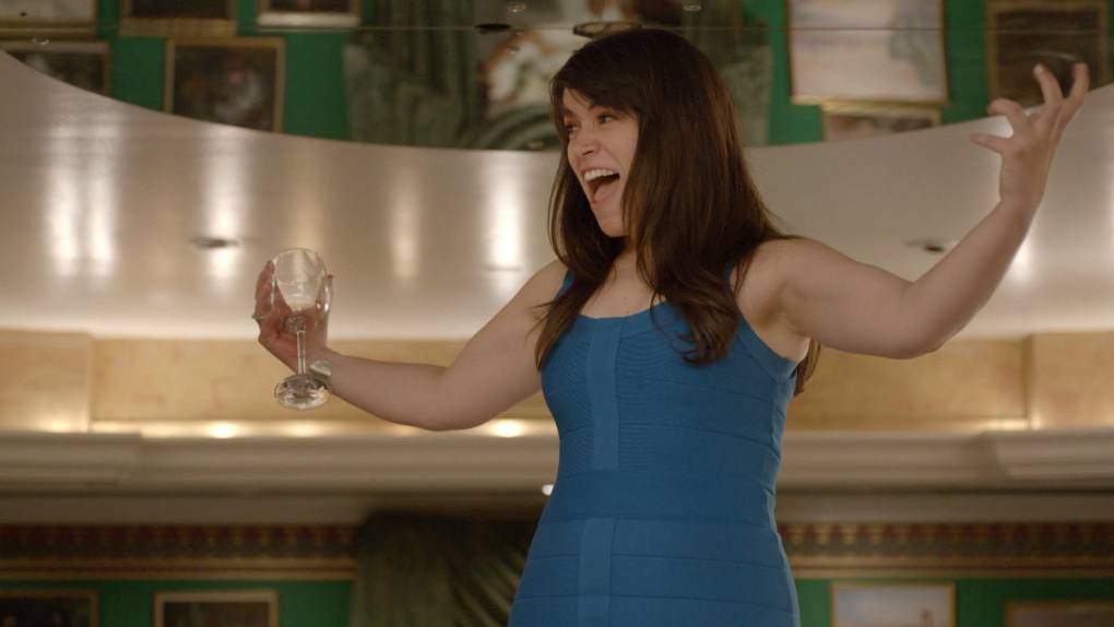 A white woman with long dark brown hair, wearing a blue dress, holding an empty glass of wine and screaming, arms outstretched.