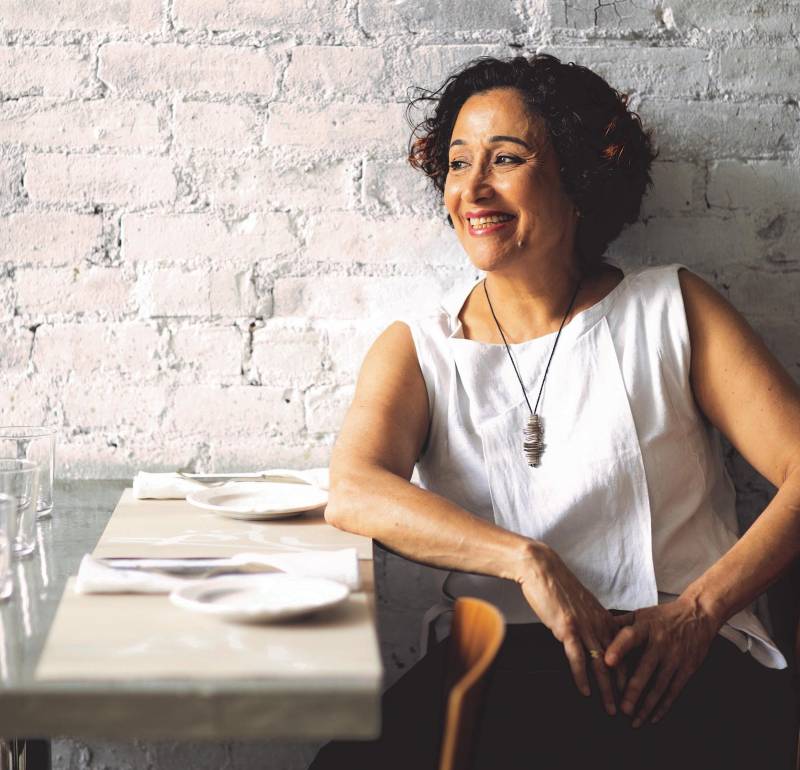 A woman of color wearing a white top sits in front of a white wall at a table. She's smiling.