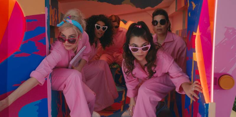 Four women in pink jumpsuits and sunglasses peak out of the back doors of a van. Partially obscured in the back is a man in the same pink jumpsuit.
