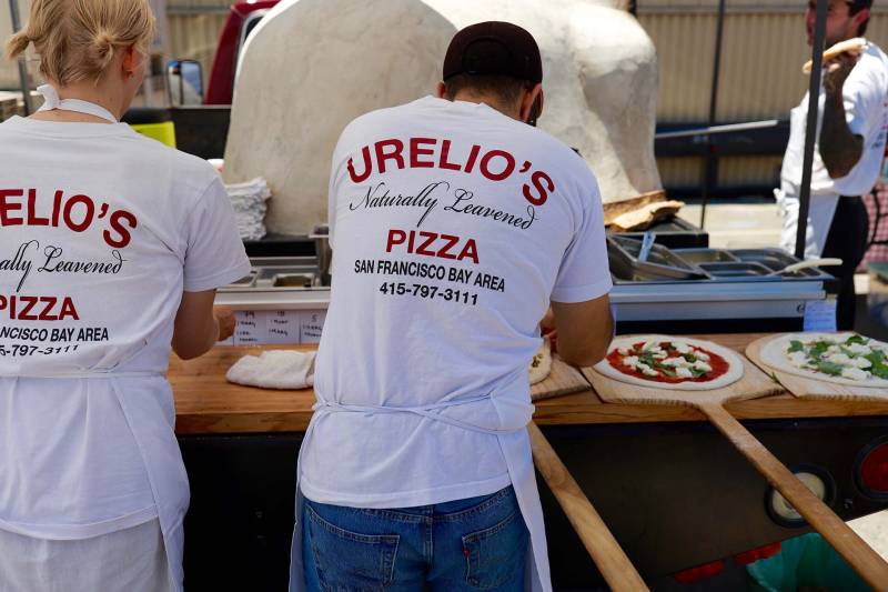 A man wearing a Urelio's Pizza shirt places toppings on pizzas at an outdoor food pop-up in Berkeley