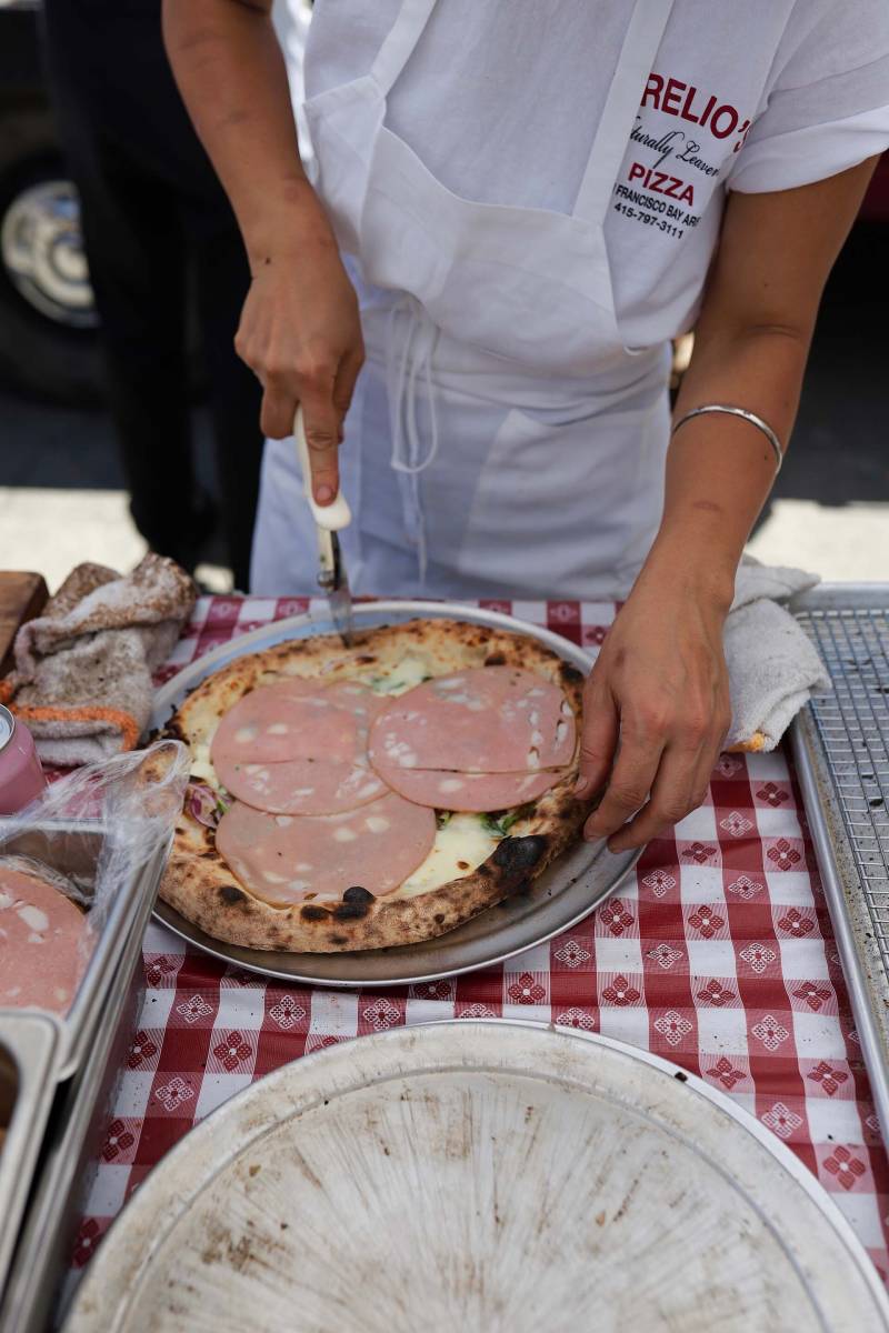 A woman wearing a Urelio's Pizza shirt slices a baloney and cheese pizza at an outdoor food pop-up in Berkeley