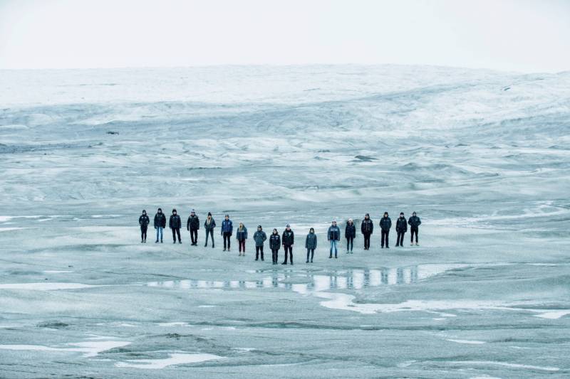 A line of people stand in a V formation atop an icy surface.