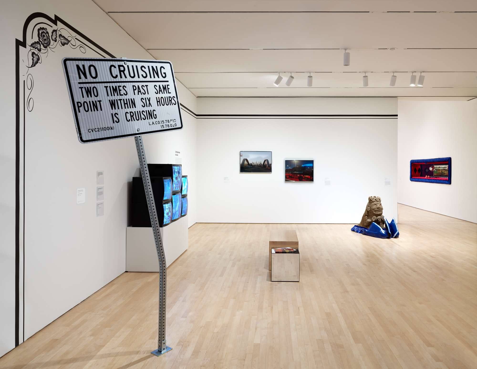 View of white-walled gallery with road sign in foreground, stacked monitors on left, photos behind and a sculpture on the floor