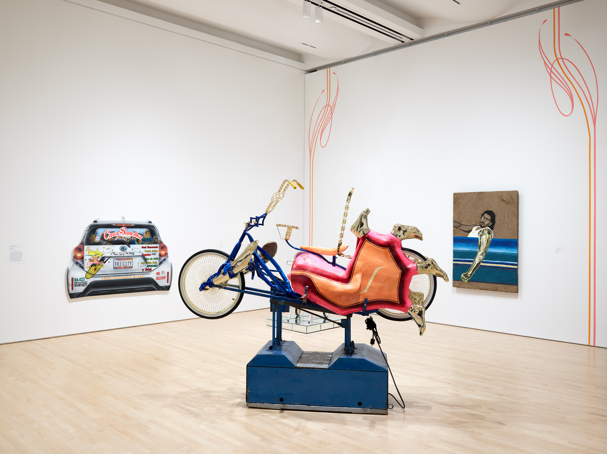 White gallery with two painting hanging on walls, one car-shaped, and a mechanical sculpture in the middle