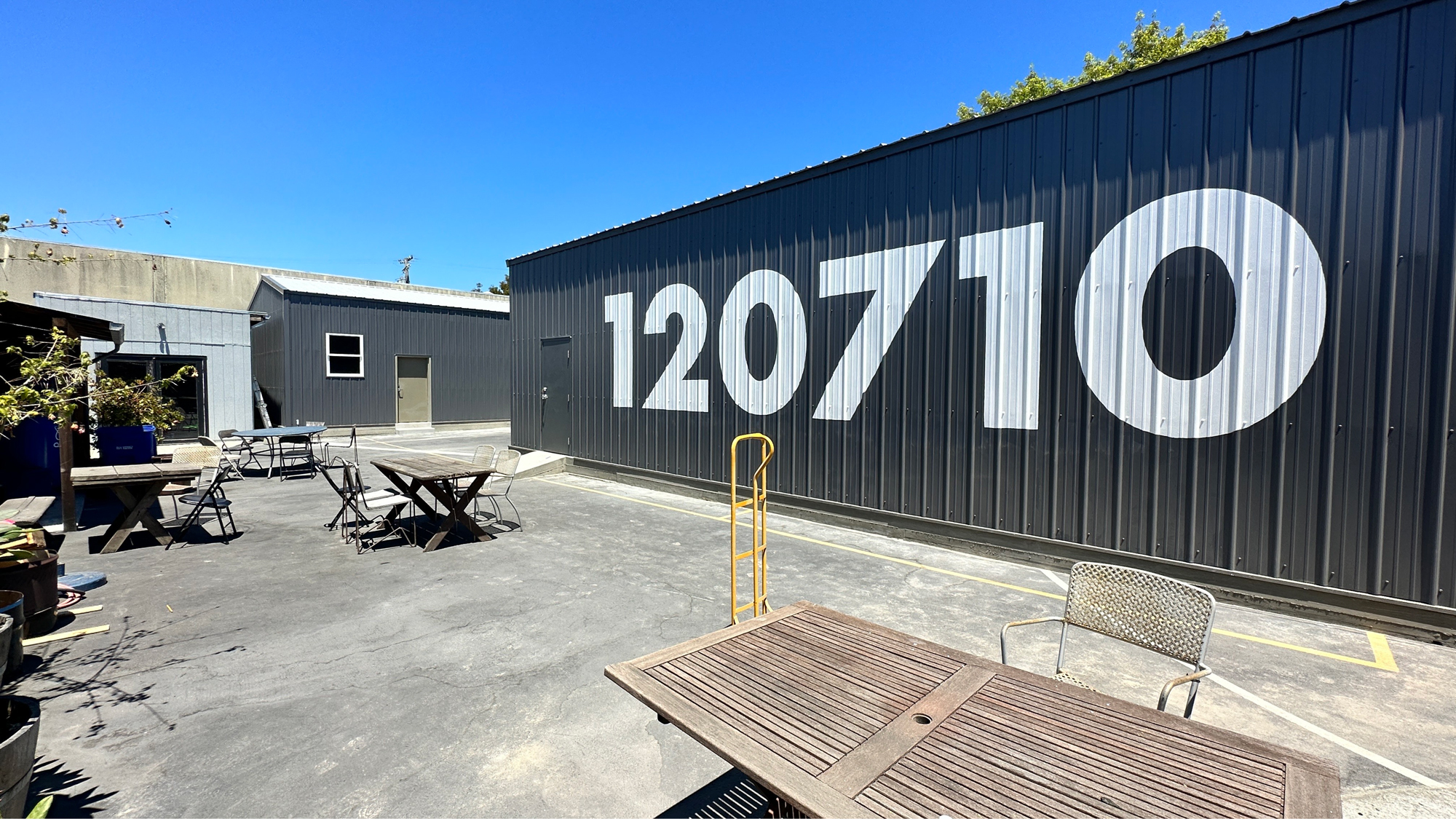 Concrete courtyard with tables bounded by gray containers, one with "120710" painted on the side in large white numbers