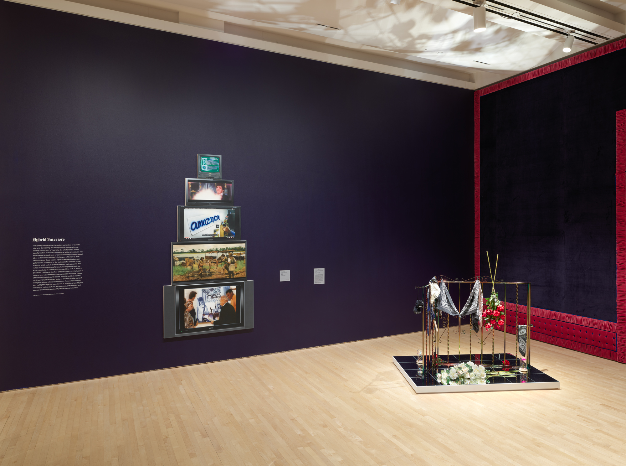 A purple-walled gallery with a stack of paintings on left wall and a highly reflective mirror-based sculpture on the right
