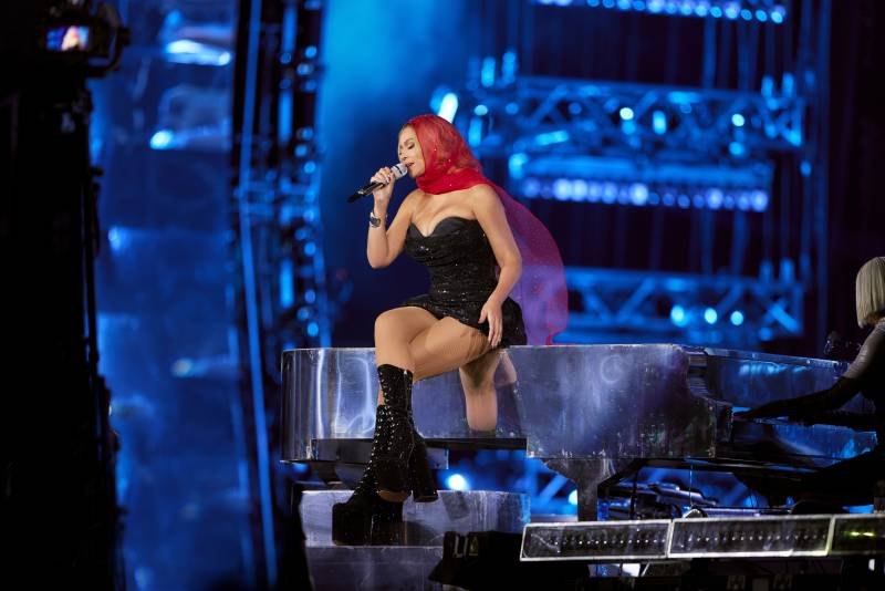A Black woman in red headpiece and black dress and tall black boots sings sitting on a grand piano