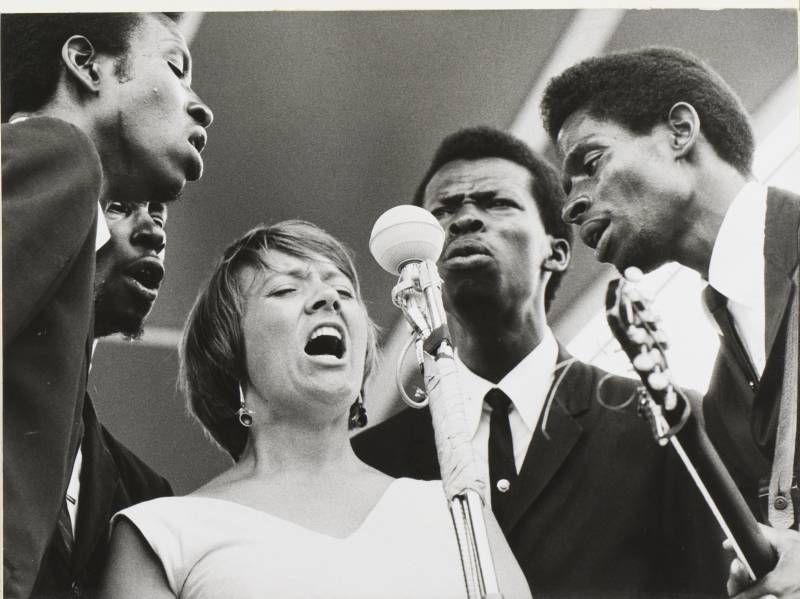 a group of singers around a microphone, four Black men and one white woman, in a black and white photo