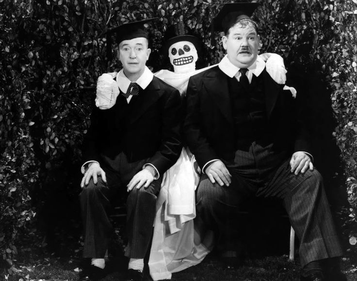 Two white men in 1930s-era suits and ties sit on the knees of a person wearing a white sheet, black hat and skeleton face paint. The men are wearing graduation caps.