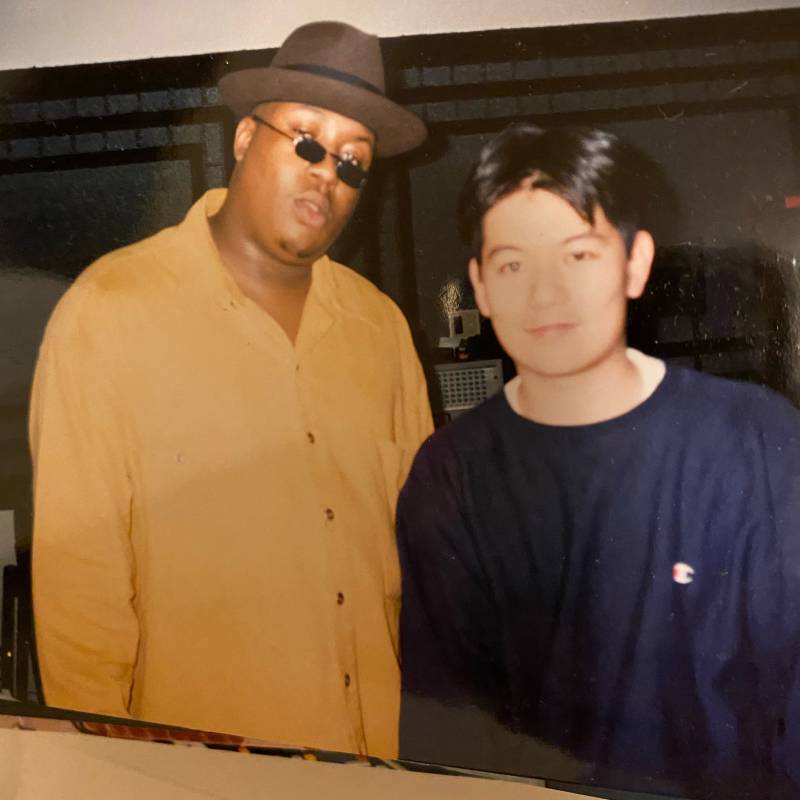 A film photo from the '90s of E-40, wearing a large hat and sunglasses, with Andy Kawanami, who wears a crewneck and smiles. 