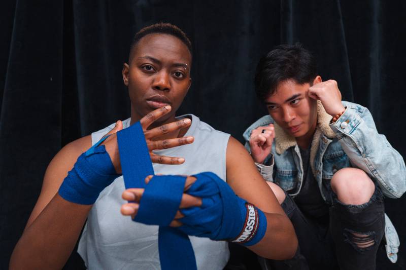 a Black woman with short hair wraps her hands in tape like she's about to box next to a young Asian American man in a jean jacket with his fists up