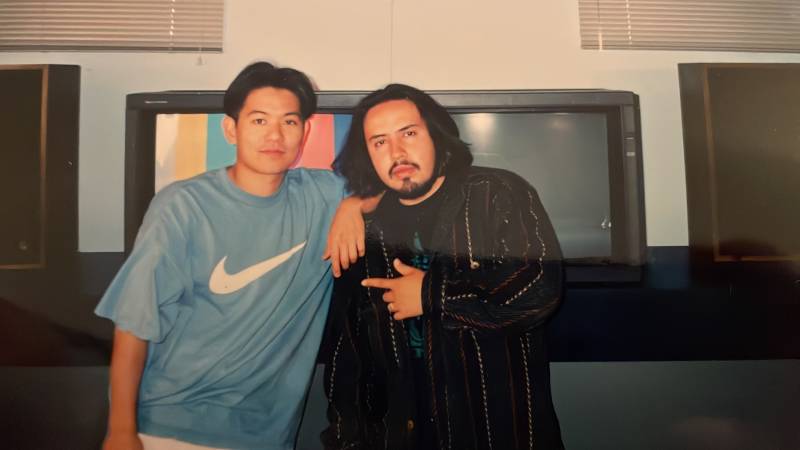 An old film photo of an Asian American young man and a Mexican American young man behind the scenes in a television studio.