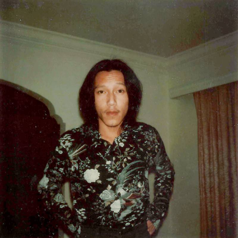 A young, slender Chinese American man with chin length hair stands in a living room wearing a black and white floral shirt, his hands in his pockets.