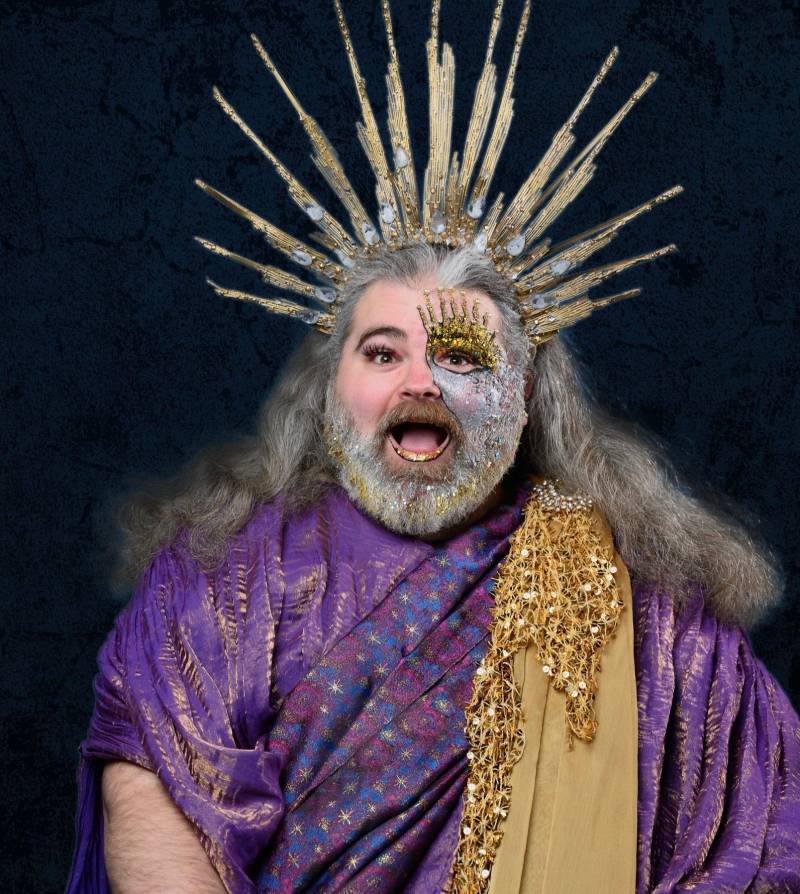 A man in purple top and spiked headpiece, with gold sash