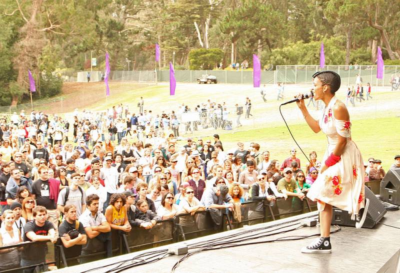 a young Black woman in a white and red dress and black and white sneakers performs onstage at a music festival in Golden Gate Park