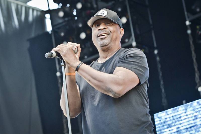 A middle aged Black man clutches a microphone and mic stand and smiles on stage. He is wearing a black t-shirt and black baseball cap.