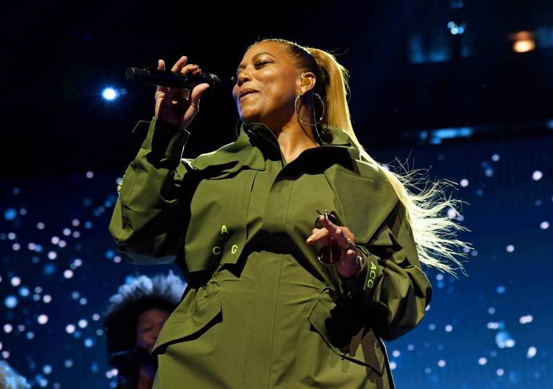 A Black woman in an olive green jumpsuit performs on stage. She is holding a microphone up to her mouth.