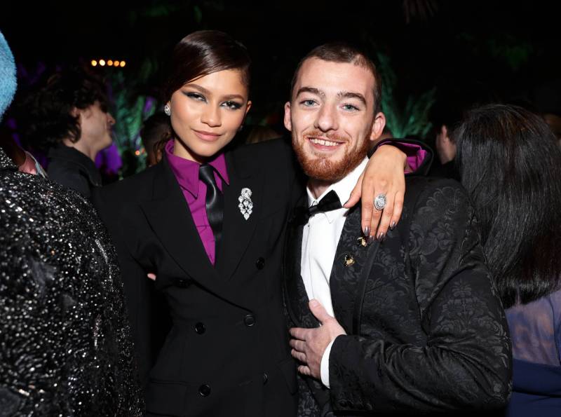 A young mixed race woman wearing a black suit and purple shirt drapes one arm over the shoulder of a smiling, bearded white man who is wearing a tuxedo.