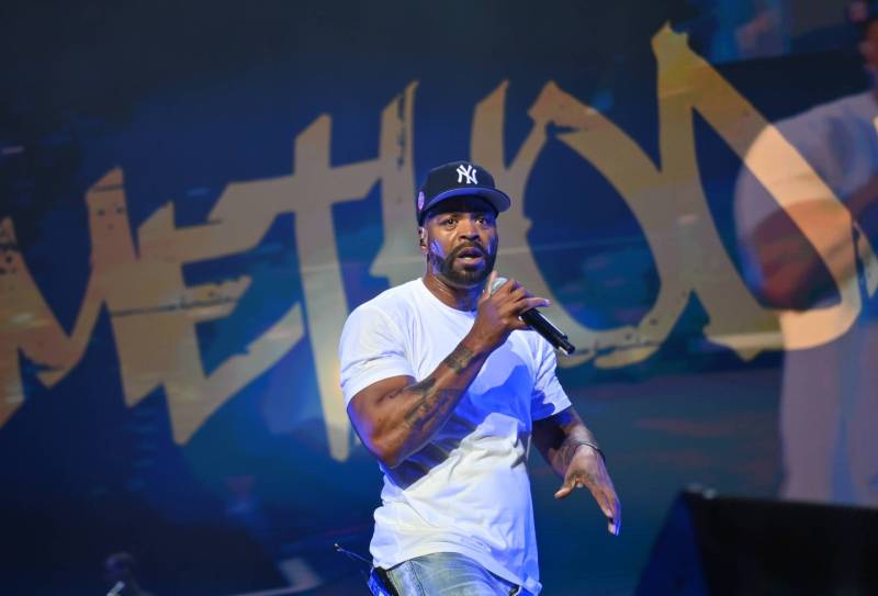 A muscular Black man, wearing an NY baseball cap, white t-shirt and blue jeans strides across a stage, microphone held up to his open mouth.