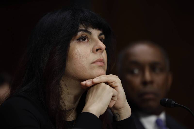A woman with pale skin and long black hair sits with her chin resting on her hands, a very serious expression on her face.