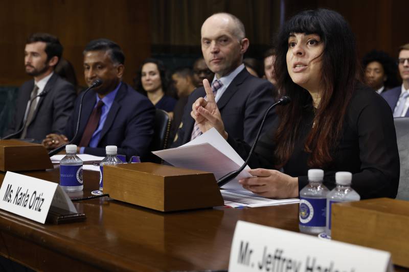A woman with long black hair points her finger for emphasis while reading a statement from white papers. In the background sit several men in suits and an audience.