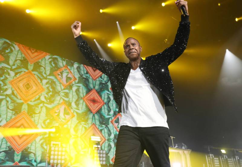 A Black man raises both his arms in the air and strides forward on stage. He is wearing a white t-shirt, black jacket and black pants. Stage lights beam all around him.