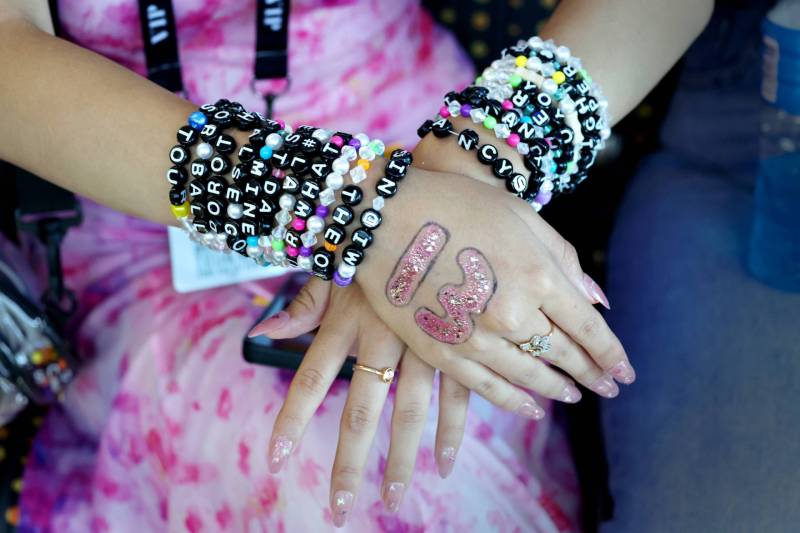 A girl's arms, loaded with inches of friendship bracelets. One hand has the number 13 drawn on it in pink glitter. The girl's pink dress is visible behind her arms.
