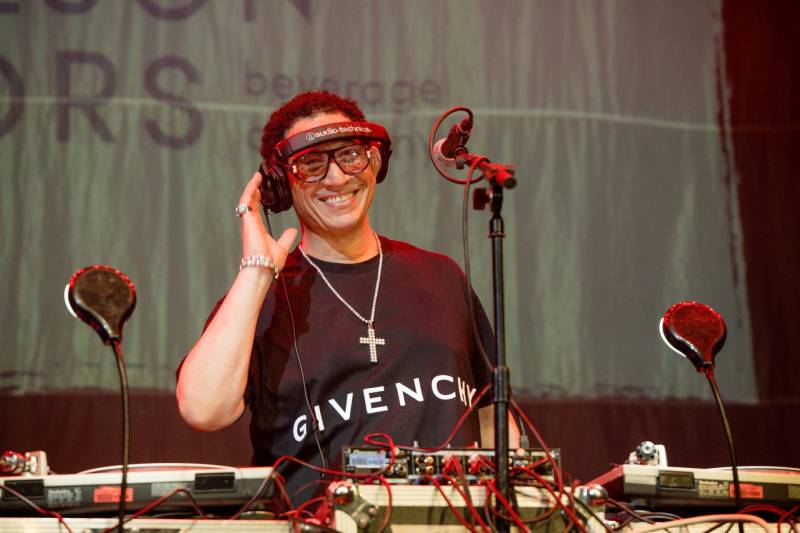 A mixed race man wearing a black Givenchy t-shirt smiles from behind DJ decks, holding one side of a set of headphones on his head.