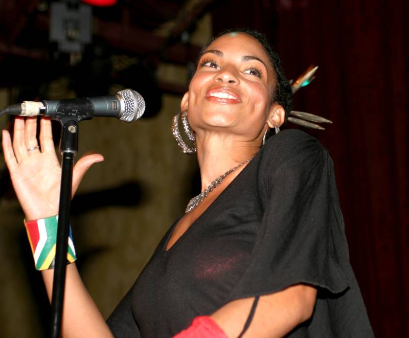 a young Black woman in a black flowy top performs while smiling at a microphone