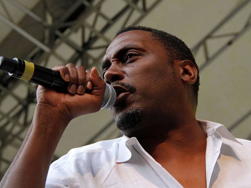 Close-up image of a Black man wearing a white shirt and rapping into a microphone on stage.