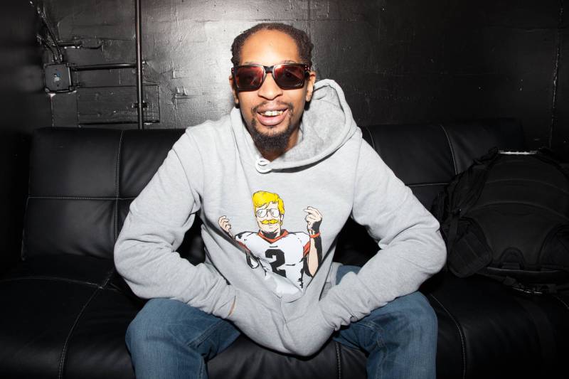 A smiling Black man wearing sunglasses, a grey hoodie and blue jeans sits on a couch next to a black wall.