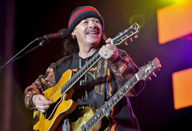 a middle-aged Latino man, Carlos Santana, smiles while playing guitar