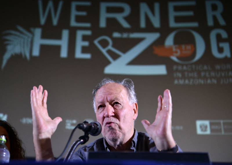 an older white man, filmmaker Werner Herzog, gestures in front of a microphone on stage