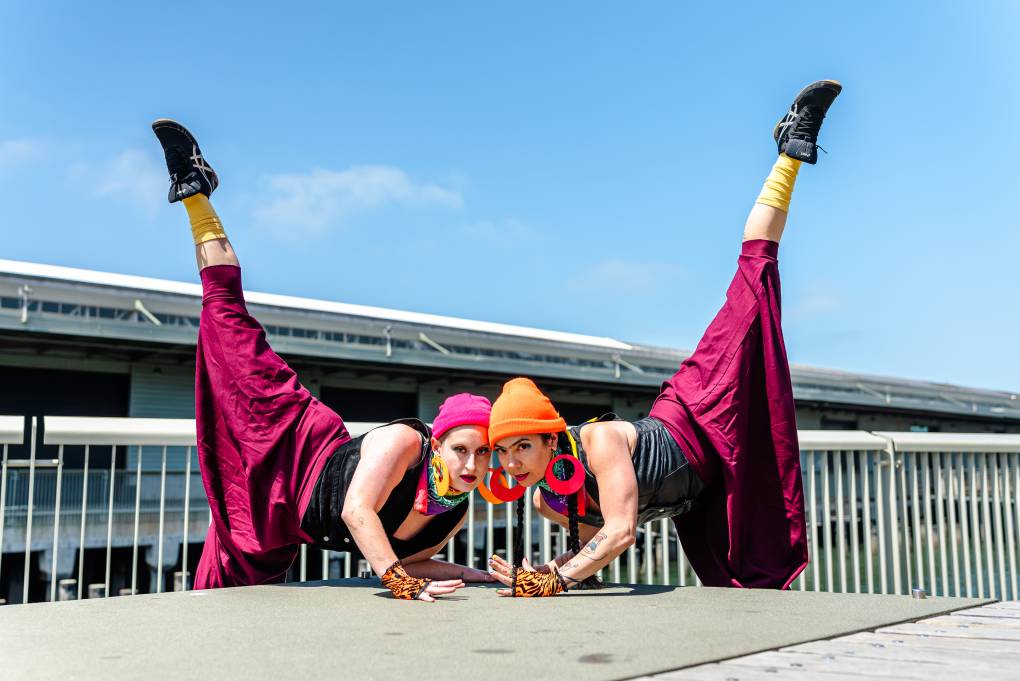 two dancers in pink and orange hat with pink skirts dance with their legs in the air, outside