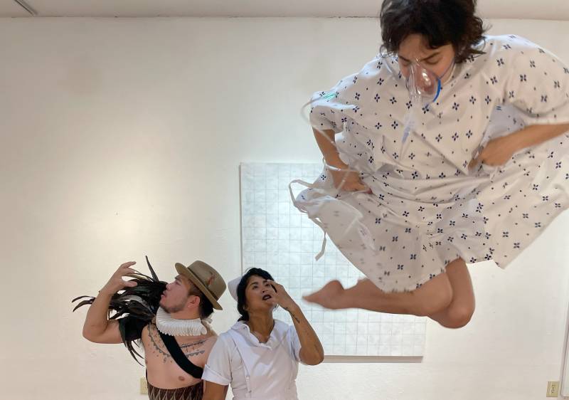 A dancer in hospital patient garb leaps in the air with a nurse in the background.